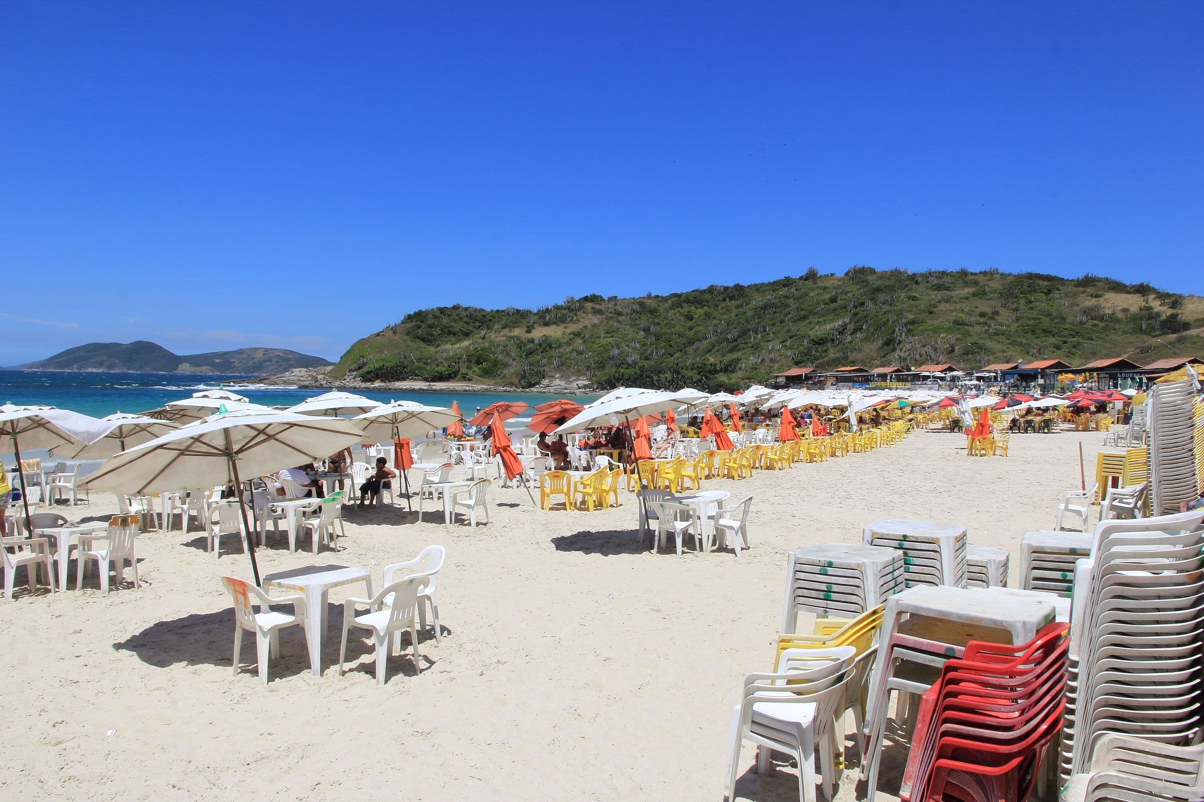 Areia clara e guarda-sóis na Praia das Conchas, em Cabo Frio, com morro verde ao fundo